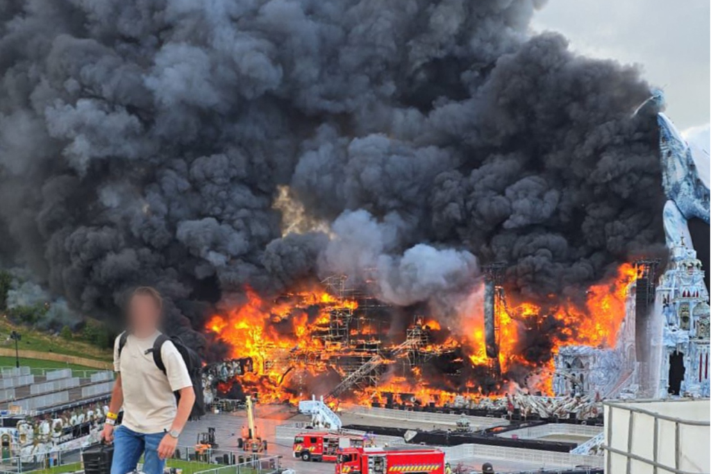 Dramatic image showing Tomorrowland 2025's Orbyz main stage fully engulfed in flames, thick black smoke rising into the sky over Boom, Belgium. Firefighters attempt to control the blaze.